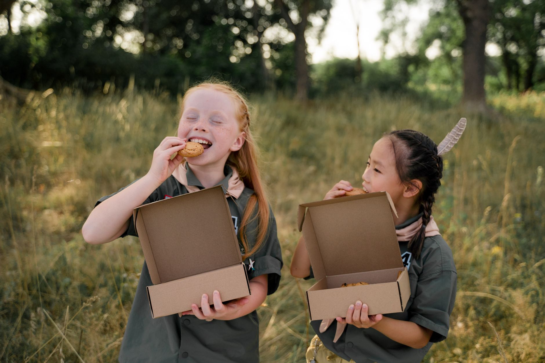 Dos chicas con uniformes de exploradoras disfrutando de galletas en un entorno natural al aire libre.