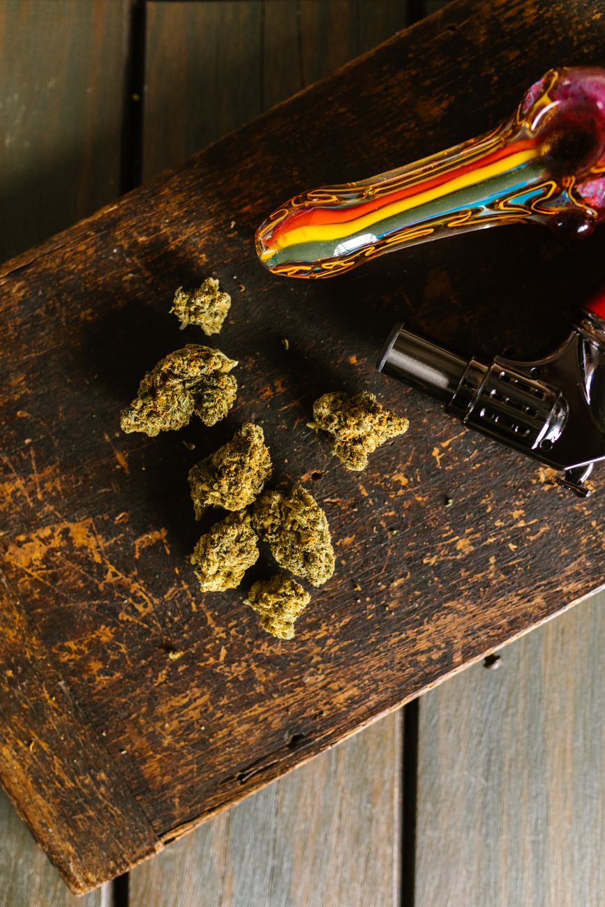 Una pipa de cristal colorida y cogollos de cannabis sobre una rústica mesa de madera en un plano vertical.