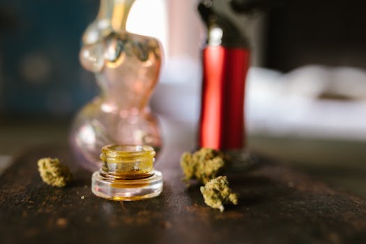 Tarro de cristal con concentrado de cannabis, rodeado de cogollos secos de marihuana y accesorios para fumar sobre una mesa.